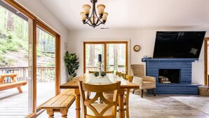 A dining area with a wooden table and chairs, a chandelier, a fireplace, and large windows overlooking a deck.