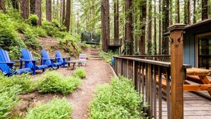 A forested deck with blue Adirondack chairs, a picnic table, and a wooden staircase leading into the woods.