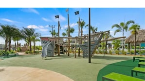 Ein Spielplatz mit Rutschen und Klettergerüsten, umgeben von Palmen und einem Gebäude mit einem Reetdach.