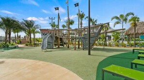 Un parque infantil con toboganes y estructuras de escalada, rodeado de palmeras y un edificio con techo de paja.