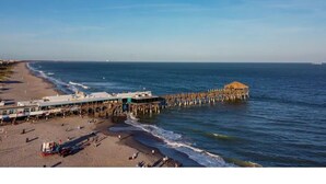 Una playa con un muelle que se adentra en el mar, un restaurante frente a la playa y una pequeña cabaña.