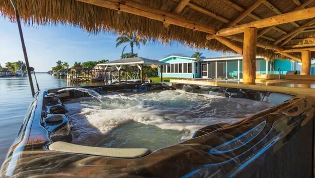 Un jacuzzi con chorros de agua, techo de paja y vista a una casa y palmeras al otro lado del agua.