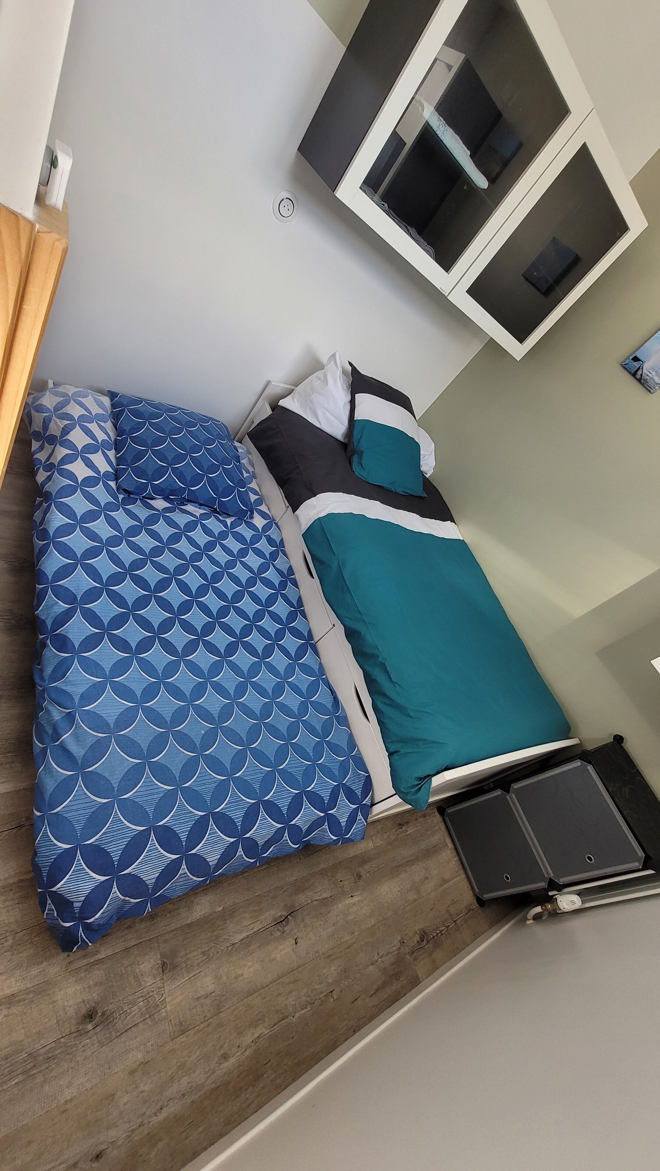 A bed with a blue patterned bedspread and teal pillows, a wooden headboard, a small bedside table with a lamp, and a wall-mounted TV.