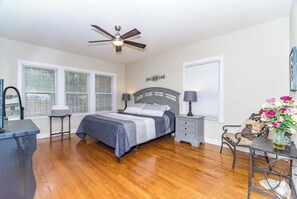 A bedroom with a bed, bedside tables, a desk, a chair, a ceiling fan, and a window with blinds.
