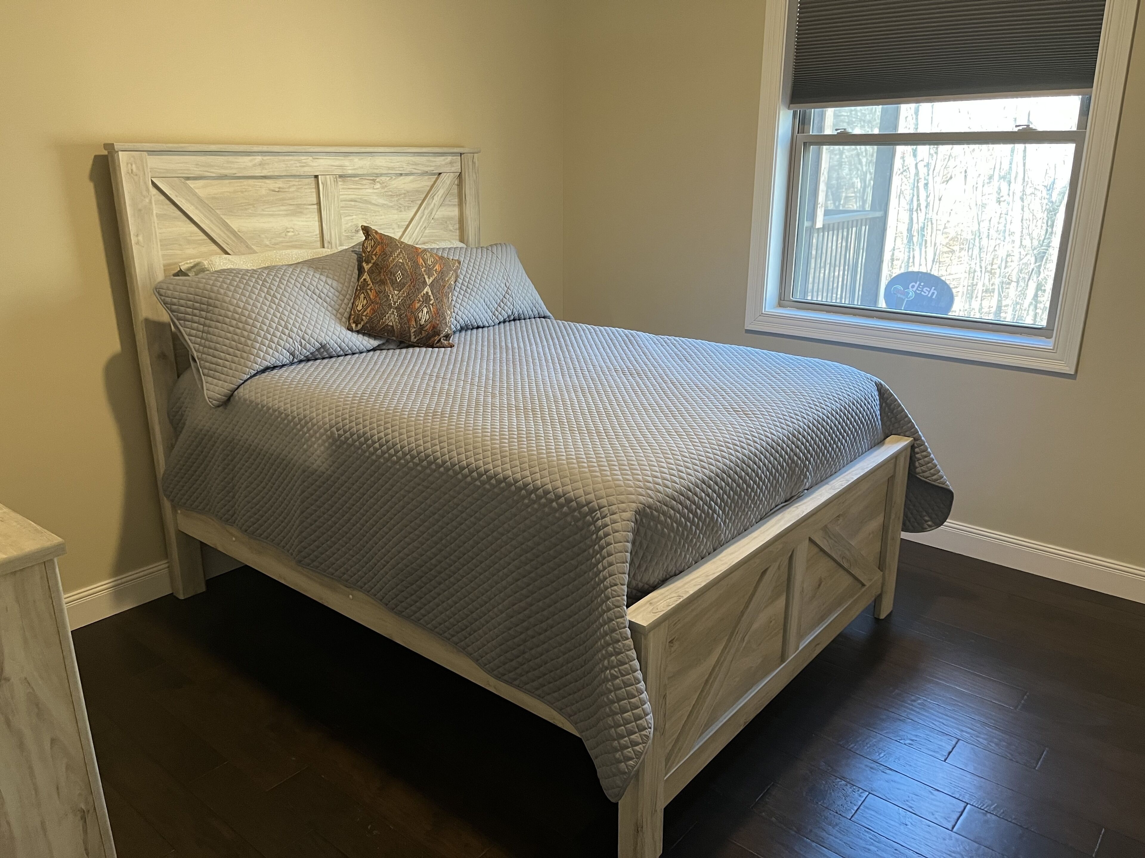 A bed with a quilted grey bedspread and a patterned pillow, a wooden headboard, and a window with blinds.