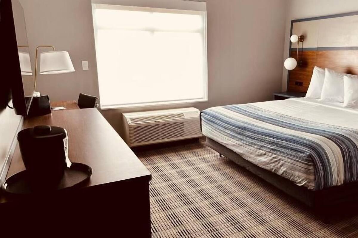 A hotel room with a bed, a desk, a chair, a lamp, a heater, and a window.