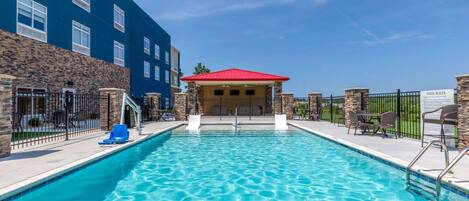 Une piscine dotée d’un pavillon au toit rouge, de murs en pierre et d’une zone délimitée par une clôture, comprenant des chaises longues.