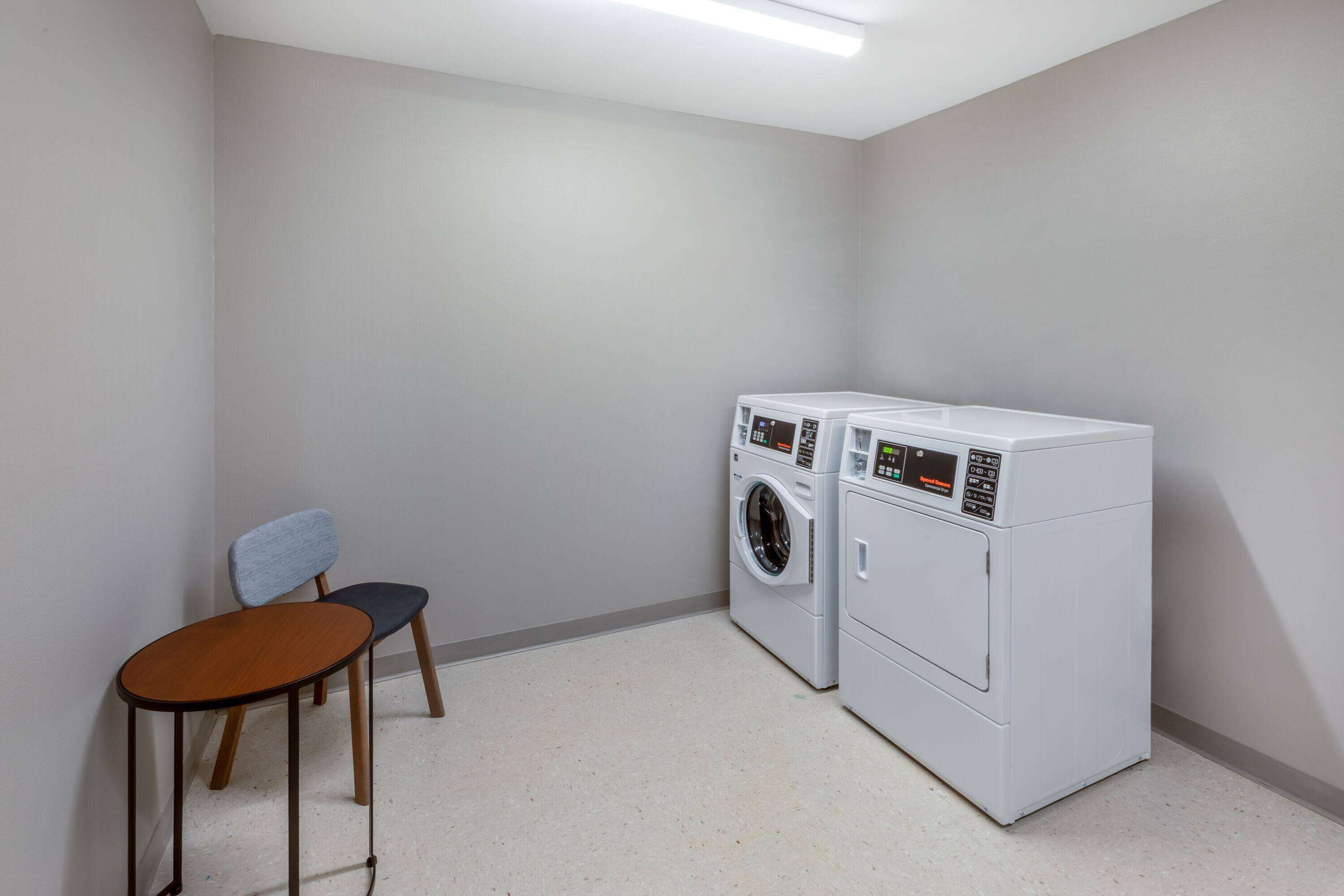 Een wasserij met twee witte wasmachines, een houten stoel en een klein tafeltje.