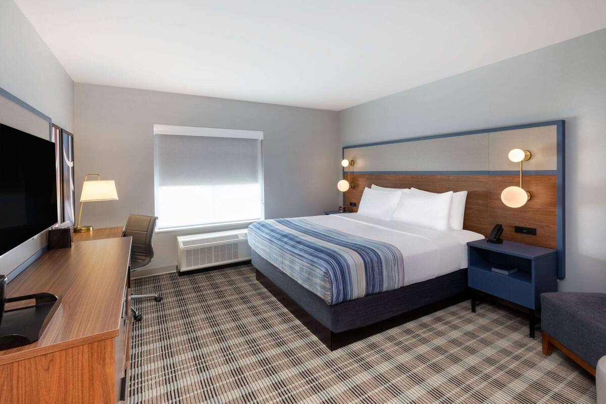 Ein Hotelzimmer mit einem großen Bett, einem Schreibtisch mit Flachbildfernseher, einem Sessel, einem Nachttisch mit Lampe und einem Fenster mit Jalousien.