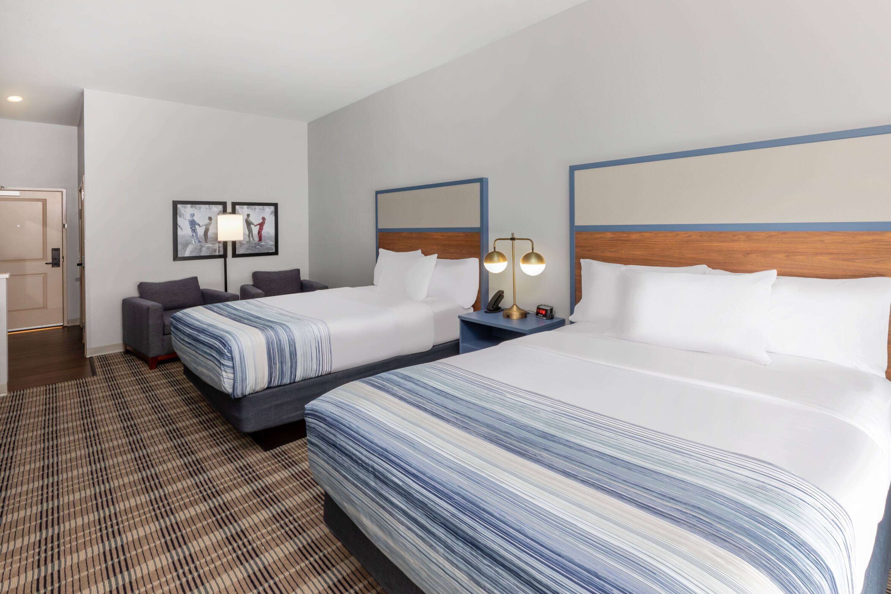A hotel room with two beds, a desk, a chair, a lamp, and a framed picture on the wall.