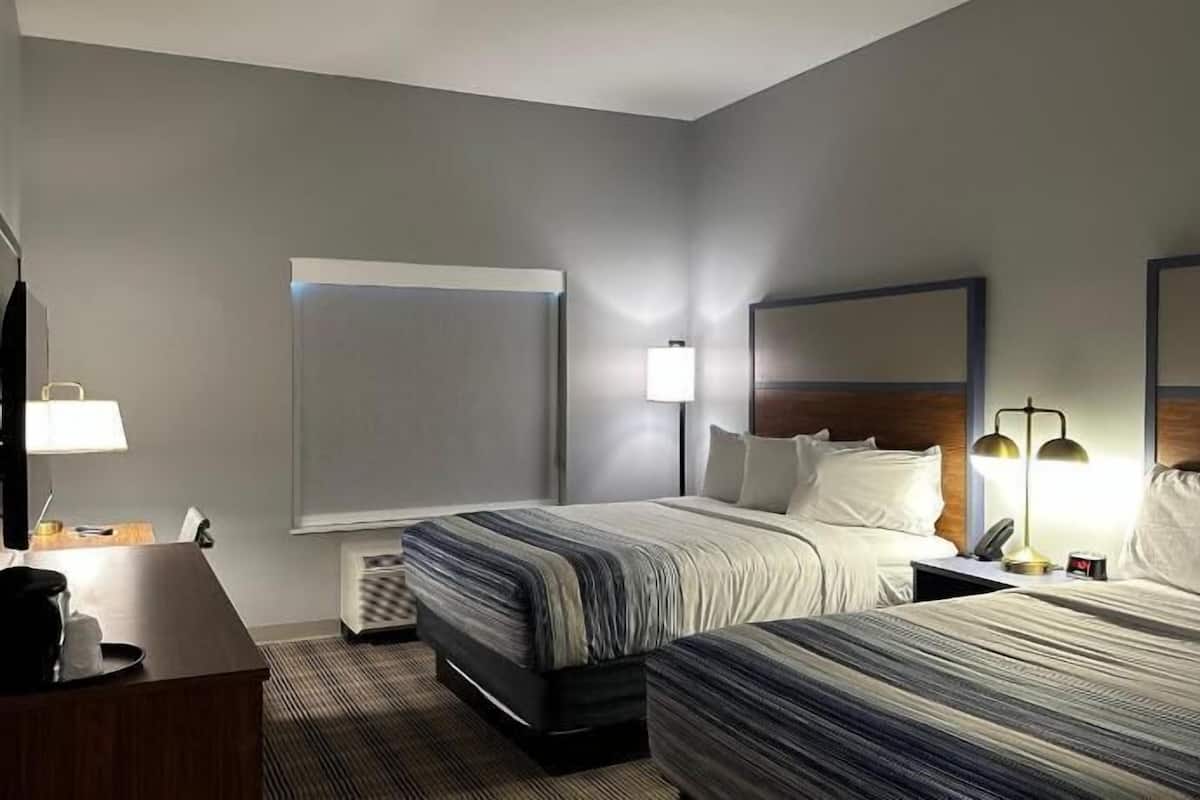 A hotel room with two beds, a desk, a chair, a TV, a lamp, and a window.
