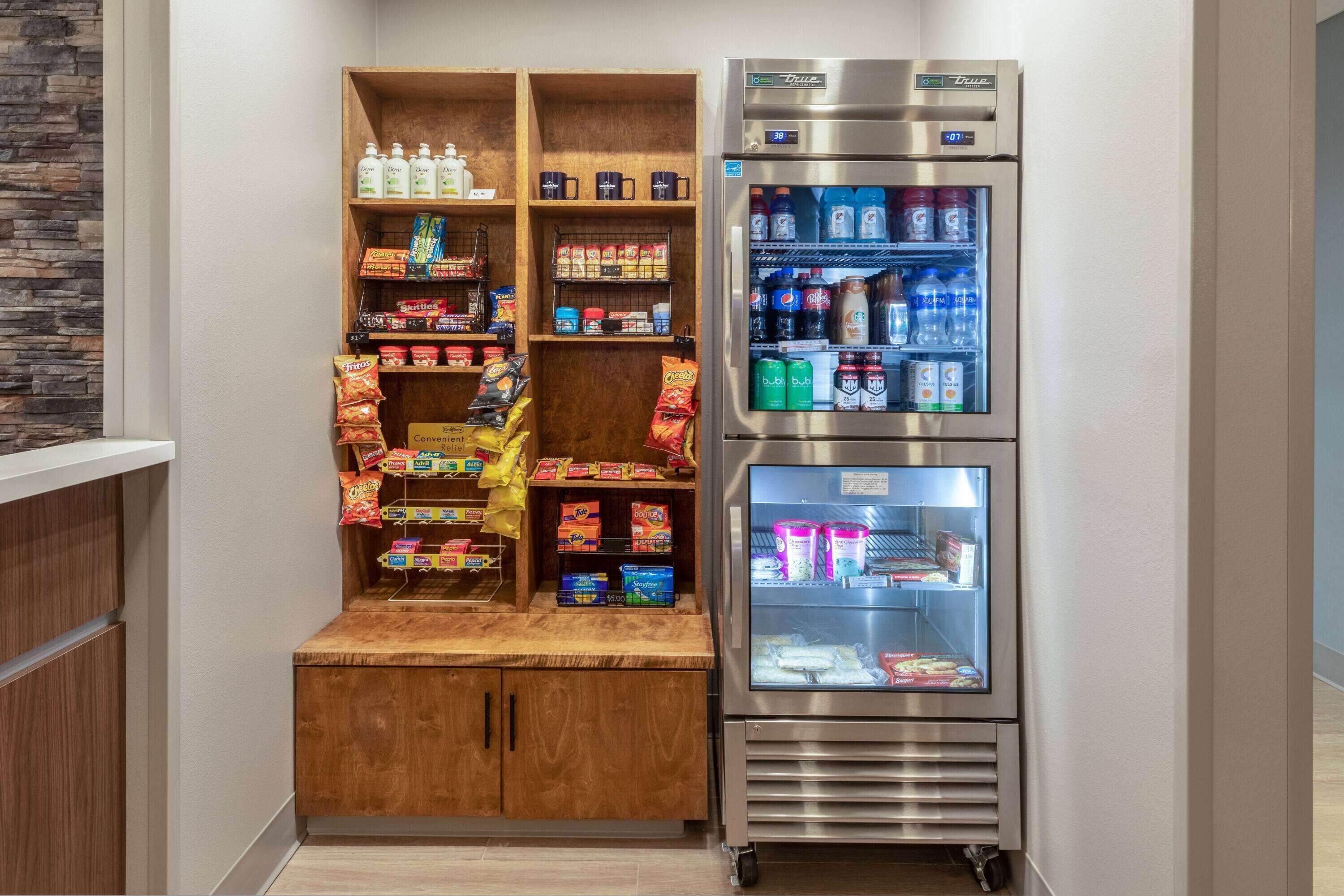 A kitchenette with a wooden shelf unit stocked with snacks and beverages, and a stainless steel refrigerator with multiple shelves.