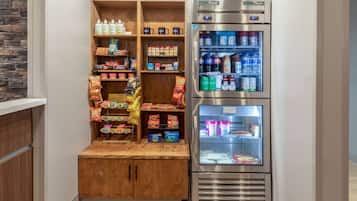 A kitchenette with a wooden shelf unit stocked with snacks and beverages, and a stainless steel refrigerator with multiple shelves.