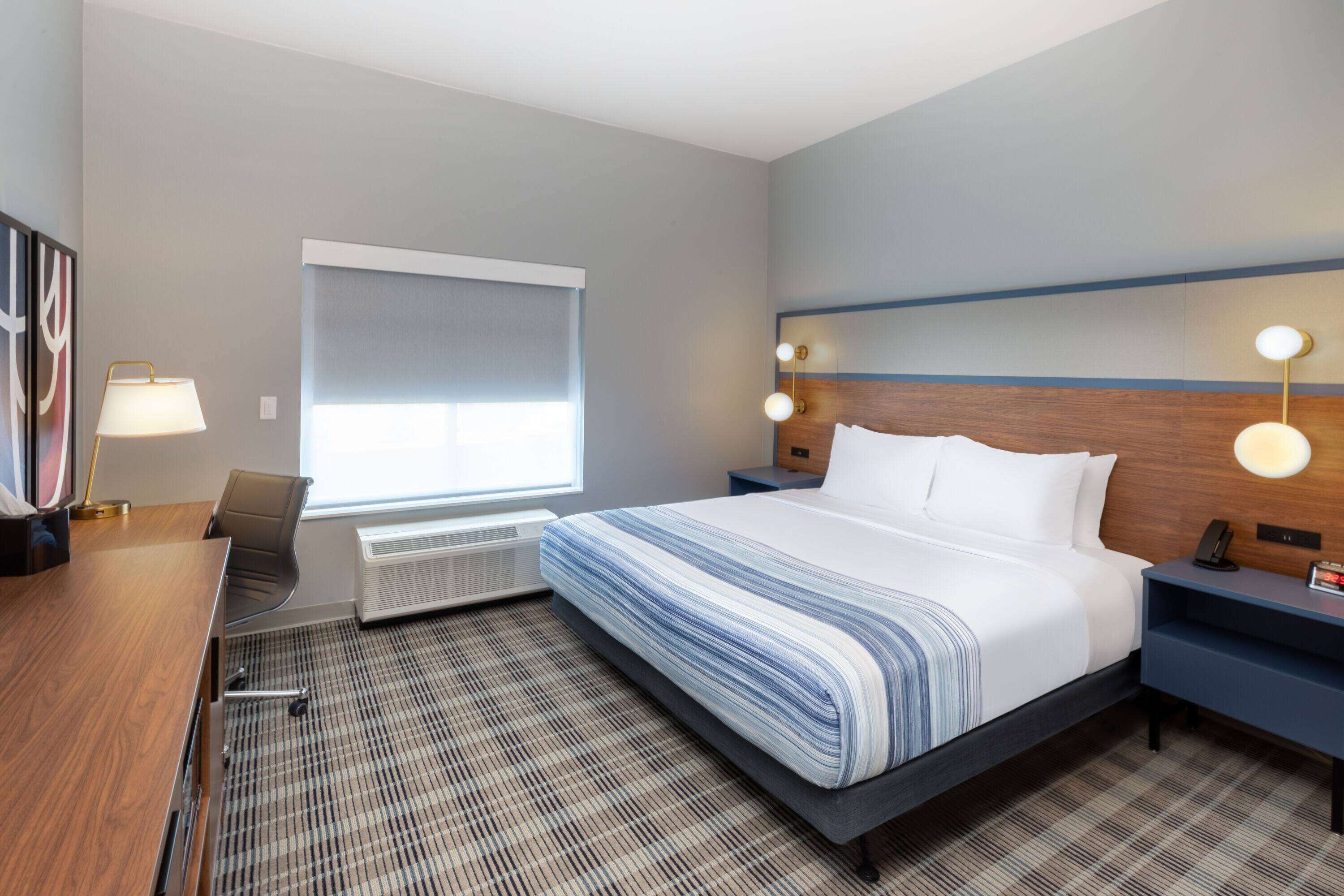 Habitación de hotel con una cama grande, un escritorio y una silla. Tiene una ventana con persianas, una lámpara de pared y un calentador.