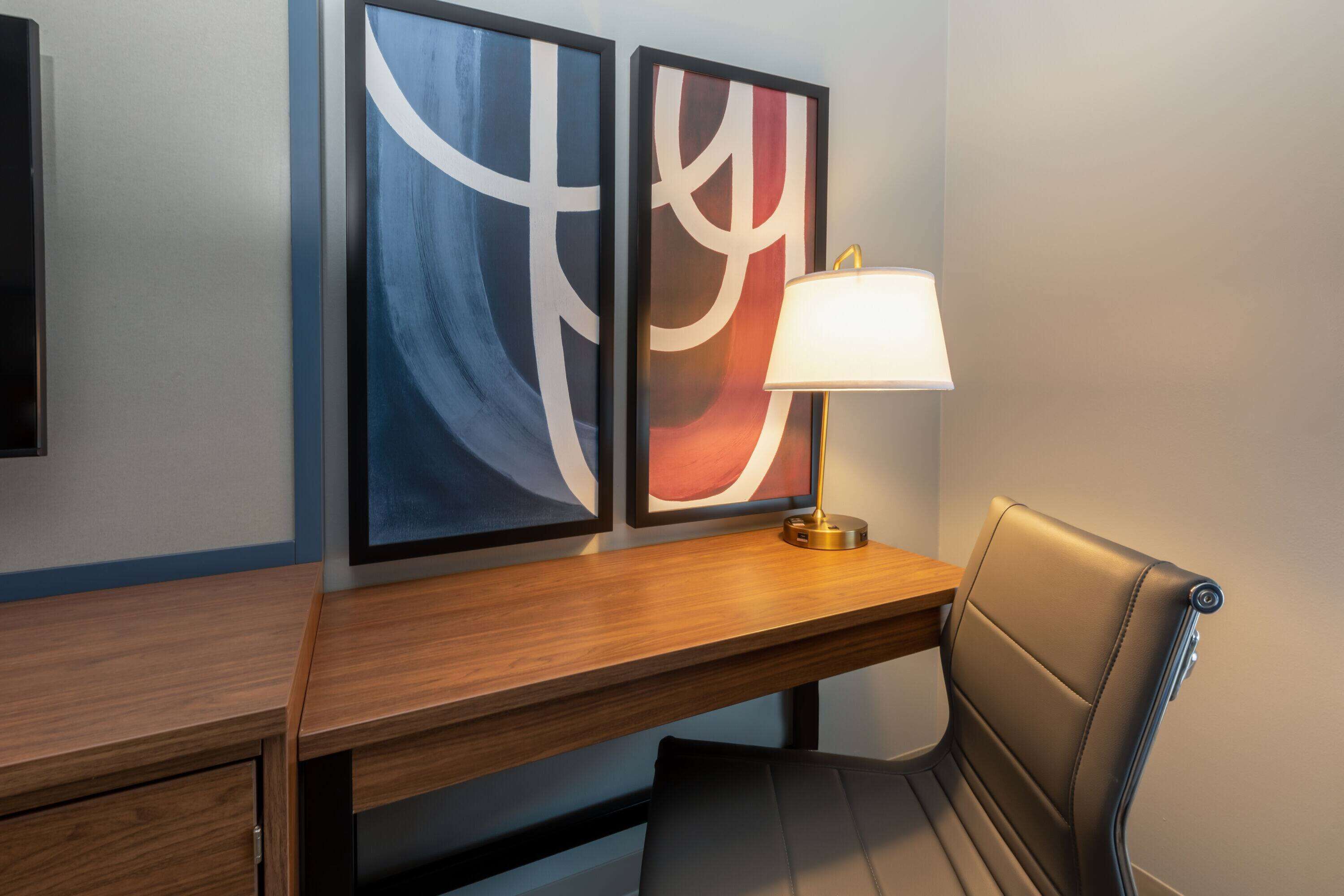 Een hotelkamer met een houten bureau, een stoel en twee abstracte schilderijen aan de muur.