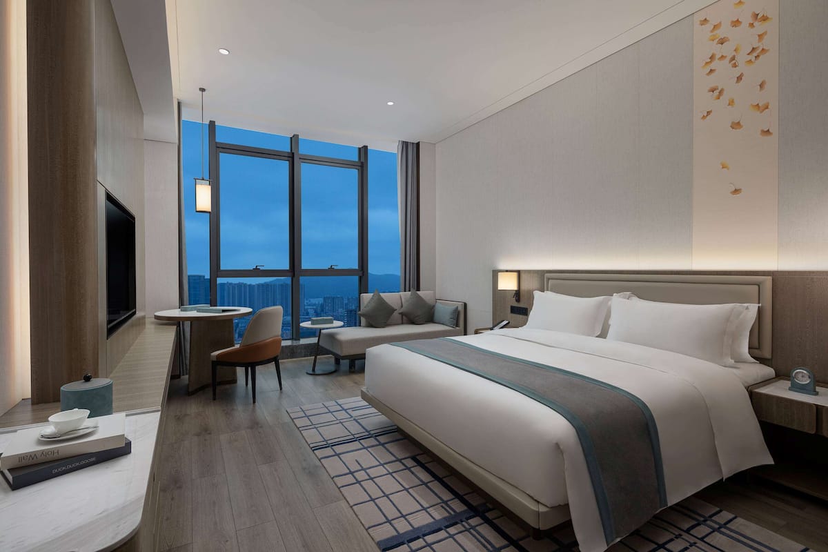A modern hotel room with a large bed, a desk, a chair, and a view of the city through large windows.