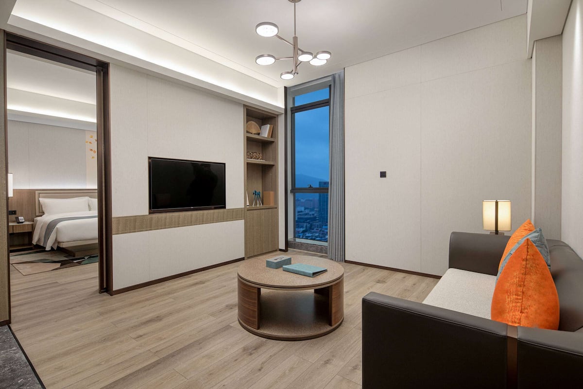 A modern hotel room with a round coffee table, a sofa, a TV, and a large window offering a city view.