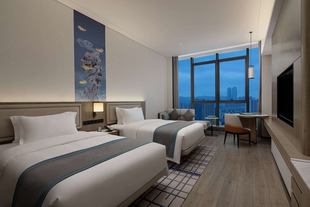 A hotel room with two beds, a desk, a chair, a TV, and a large window with a city view.