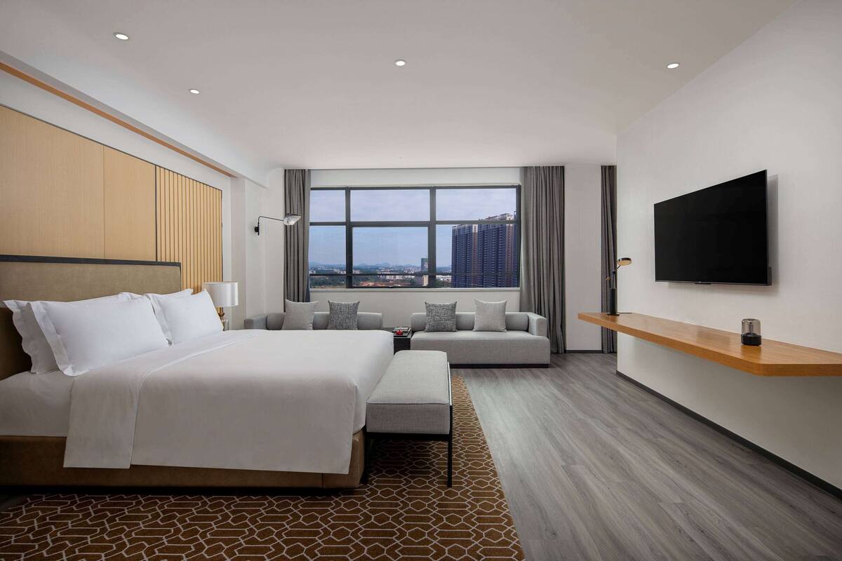 A modern hotel room with a large bed, a sofa, a TV, and a city view.