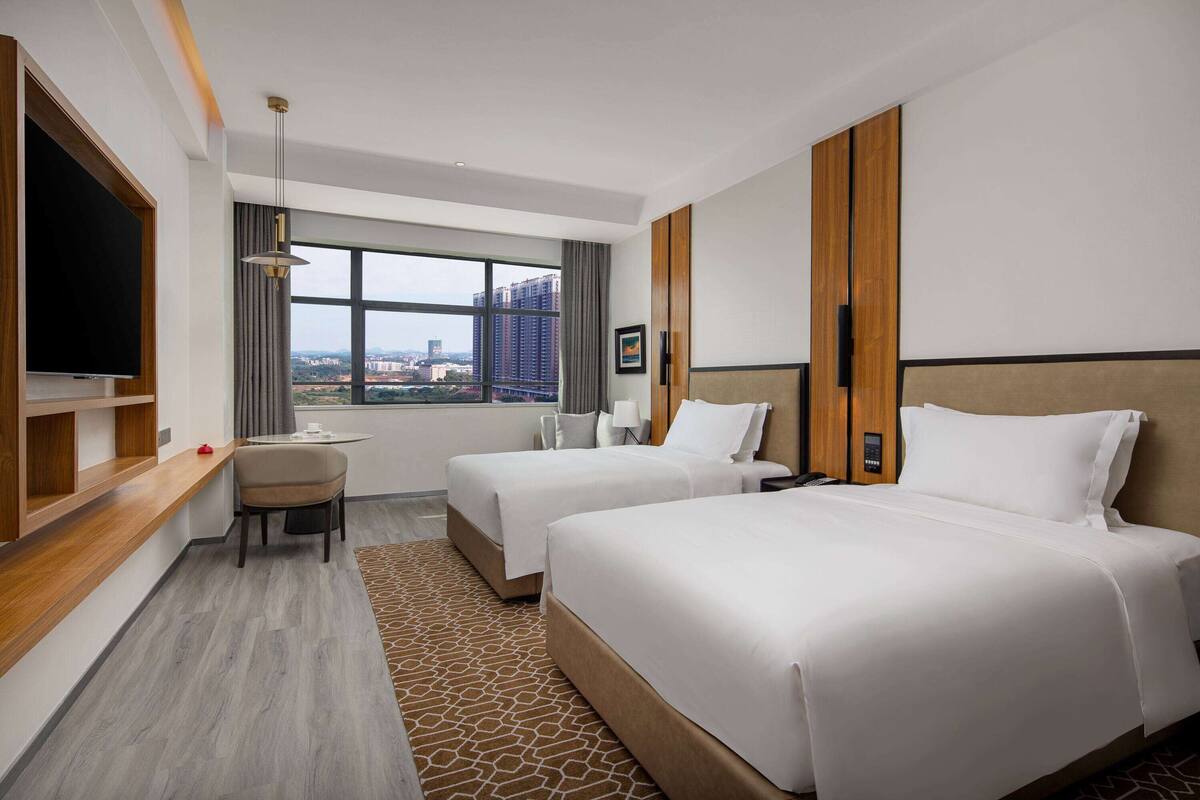 A hotel room with two beds, a TV, a desk, and a large window with a city view.