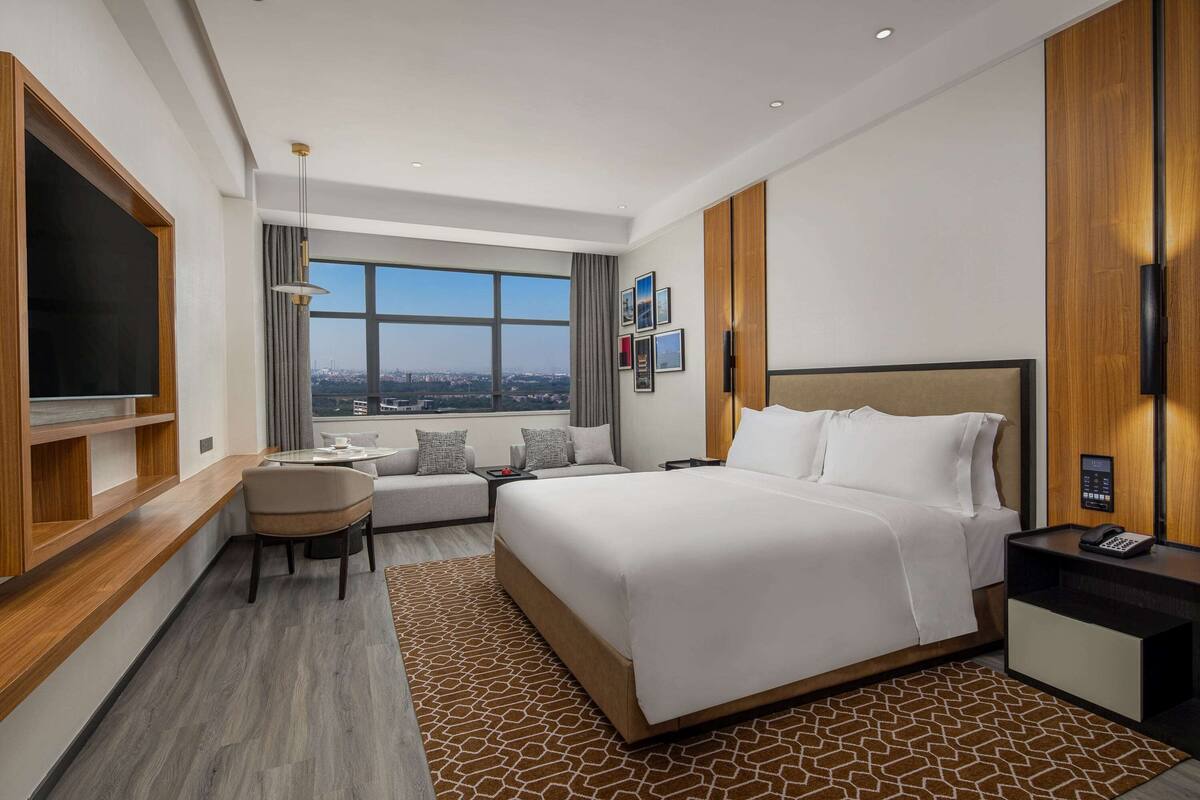 A modern hotel room with a large bed, a desk, a chair, a TV, and a view of the city.