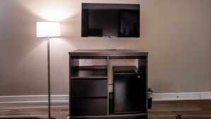 A room with a flat-screen TV mounted on the wall, a cabinet with a built-in refrigerator, and a floor lamp.
