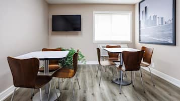 A dining area with a white table and brown chairs, a flat-screen TV on the wall, and a window with blinds.