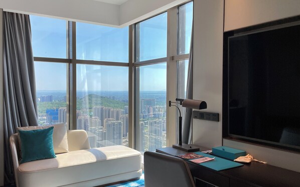 Una habitación de hotel moderna con un gran ventanal que ofrece vistas a la ciudad, un sofá cómodo con un cojín verde azulado, un escritorio con una computadora y una caja azul, y un telescopio montado.