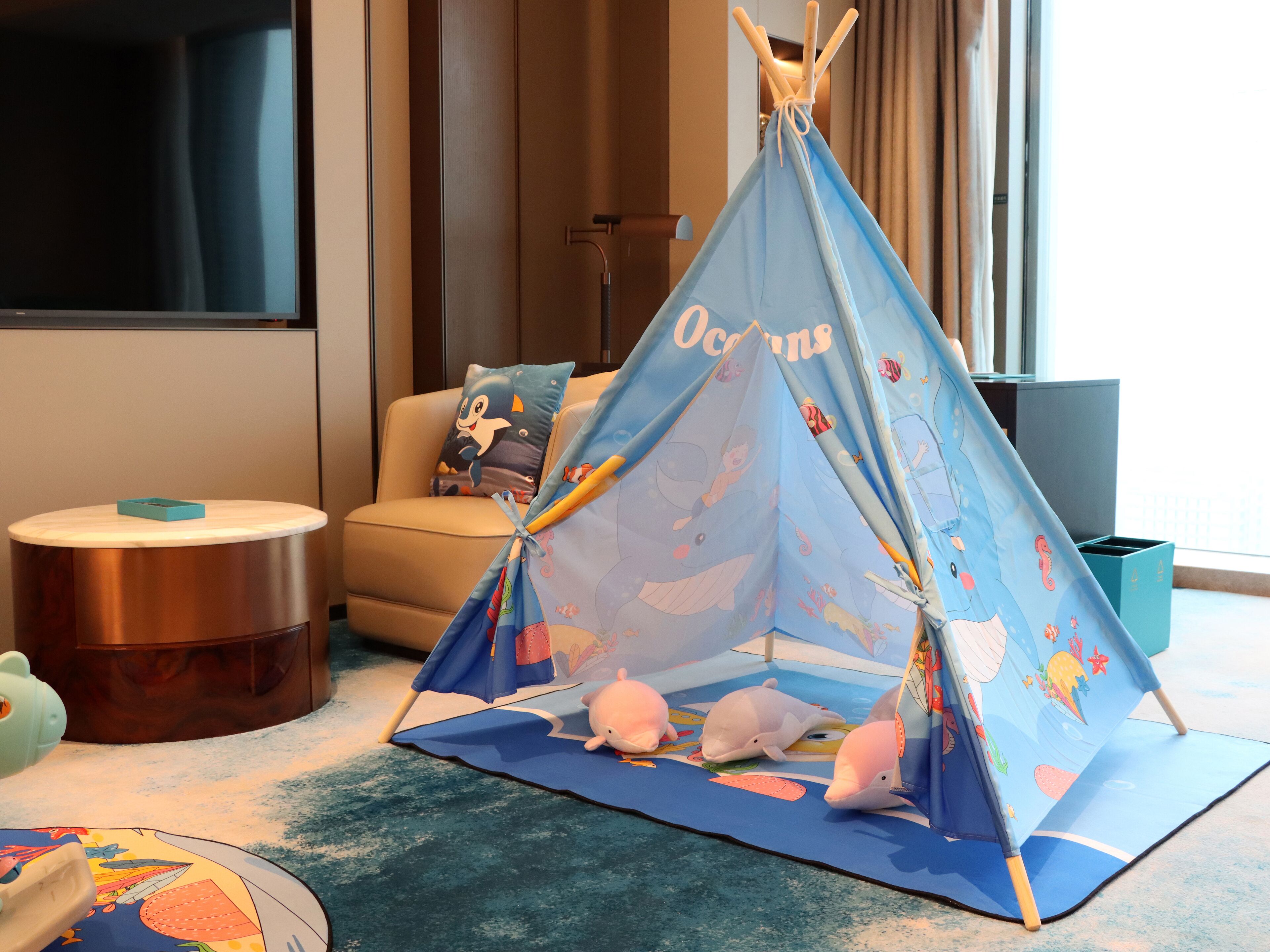 A child-friendly play area with a blue tent featuring ocean and sea creature designs, a round wooden table, a beige sofa with a penguin pillow, and a globe on the floor.