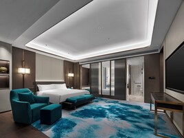 A modern hotel room with a large bed, a seating area with teal chairs, a large window, and a flat-screen TV.