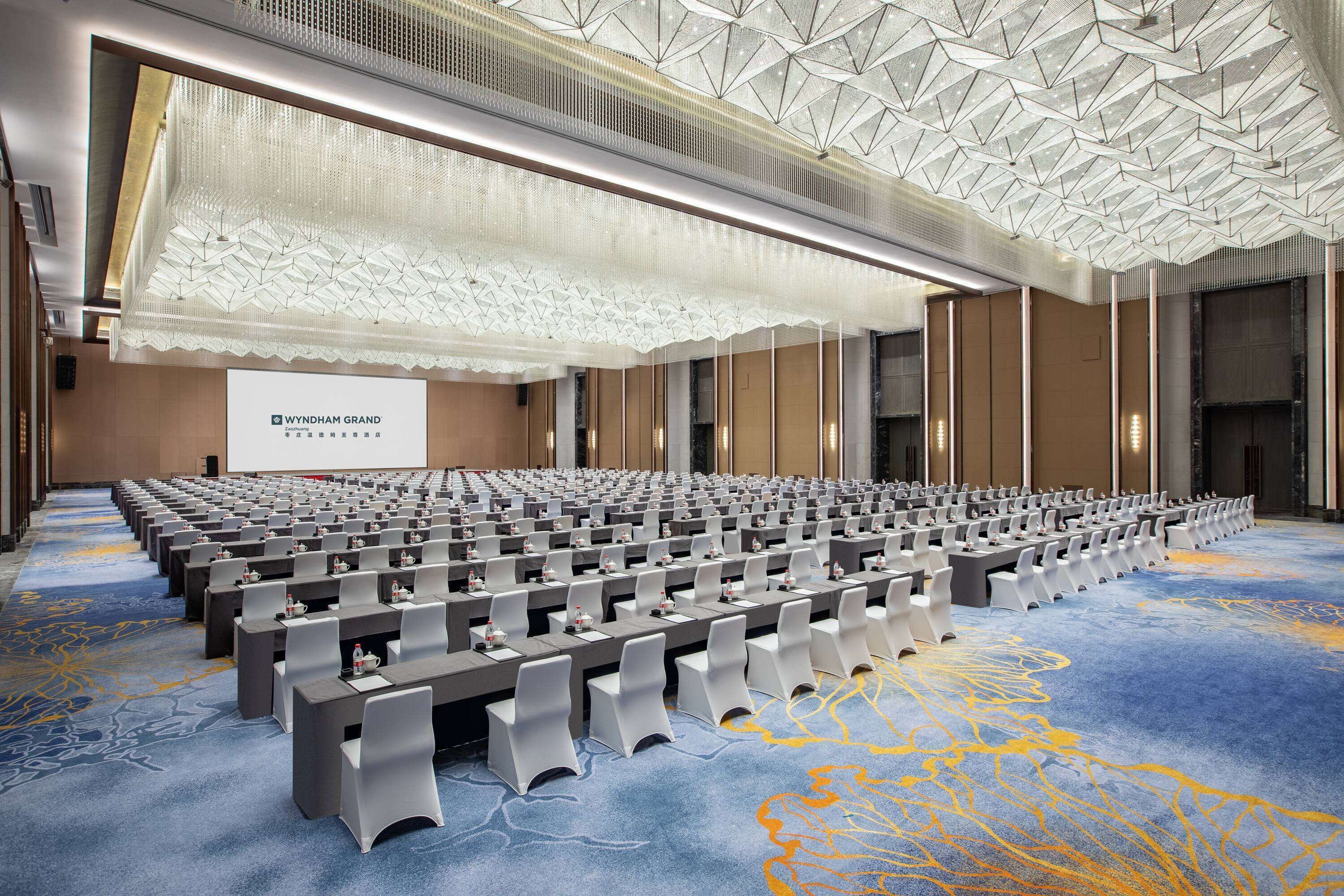 A conference room with rows of tables and chairs, a large screen, and a patterned carpet.