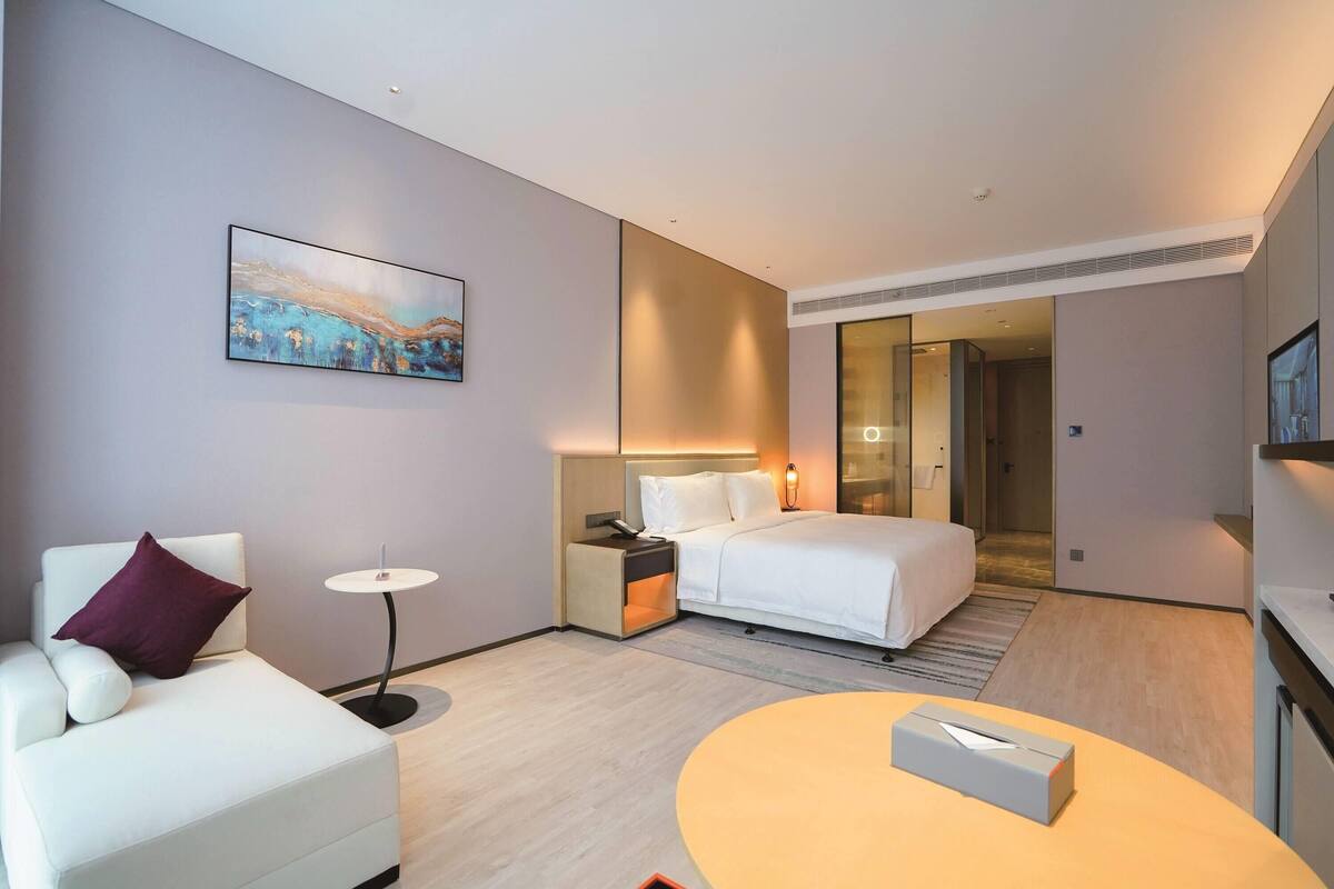 A modern hotel room with a bed, a white sofa, a small table, and a painting on the wall.