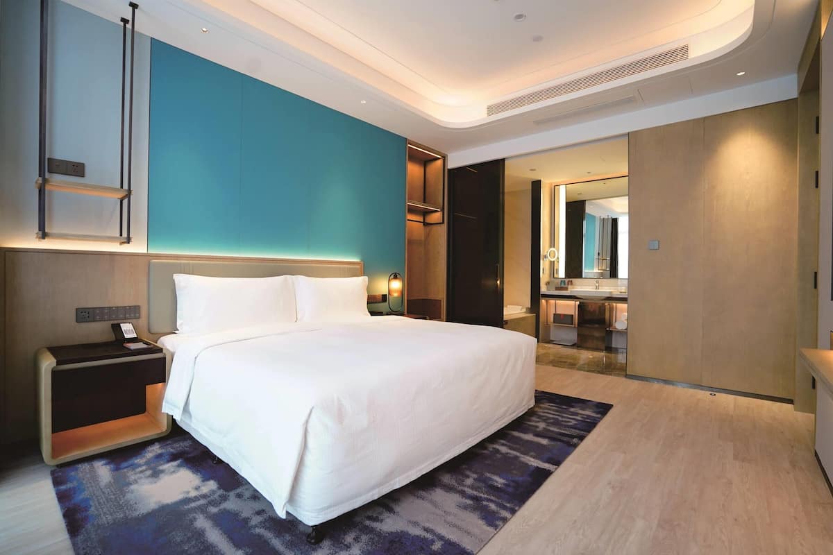 A modern hotel room with a large bed, a nightstand, a desk, and a bathroom visible through an open door.