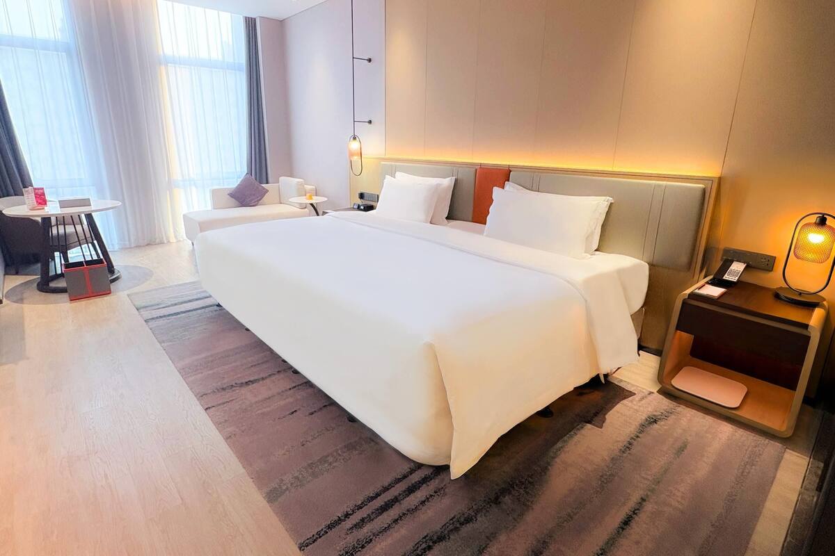 A hotel room with a large bed, a bedside table, a desk with a chair, and a window with curtains.