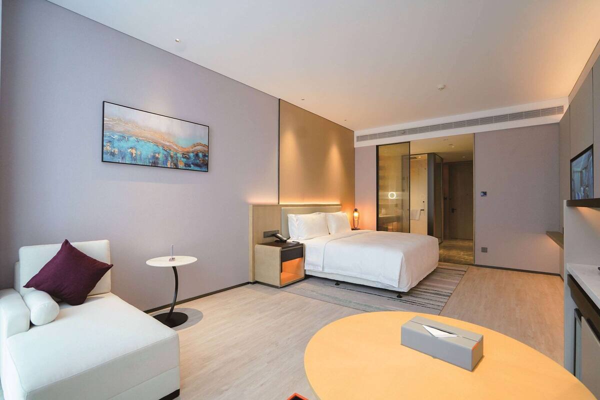 A modern hotel room with a bed, a sofa, a small table, and a painting on the wall.