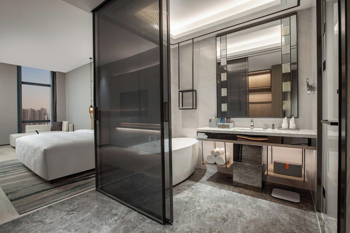 A modern hotel room with a large bed, a separate bathroom, a large mirror, and a city view.