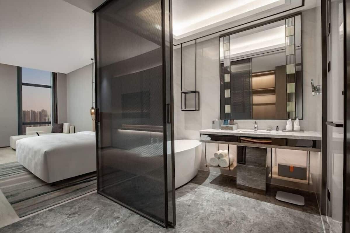 A modern hotel room with a large bed, a glass partition, a bathroom with a bathtub, and a large mirror.