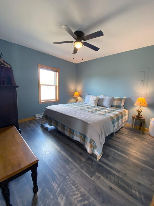 A bedroom with a bed, two bedside lamps, a ceiling fan, a window, and a wooden table.