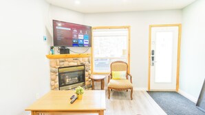 Un salon avec une cheminée, un téléviseur fixé au mur, une table basse en bois et un fauteuil confortable.