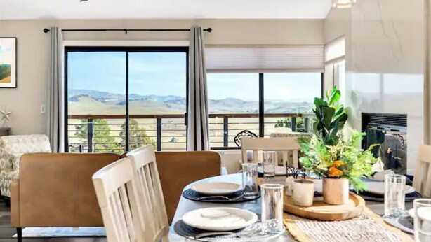 Un coin repas avec une table dressée pour deux personnes, de grandes fenêtres offrant une vue sur les collines et un ventilateur de plafond.