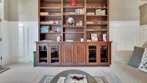 Ein Wohnzimmer mit einer Holzbücherreol, einer grünen Mülltonne und einem beigen Sofa.