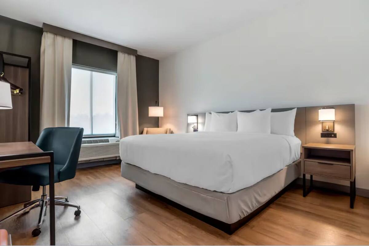 A hotel room with a large bed, a desk, a chair, and a nightstand with a lamp.