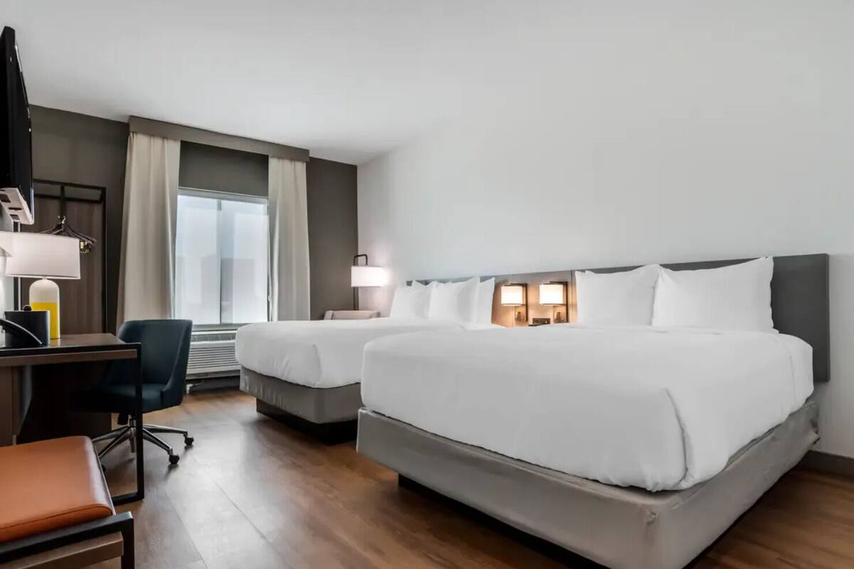 A hotel room with two beds, a desk with a chair, a lamp, and a window with curtains.