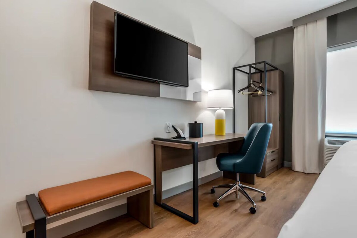 Een hotelkamer met een bed, een bureau met stoel, een aan de muur bevestigde televisie, een bedlamp en een raam met gordijnen.