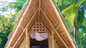 Une hutte au toit de chaume, dotée d’un lit et d’un petit espace porche, entourée de verdure.