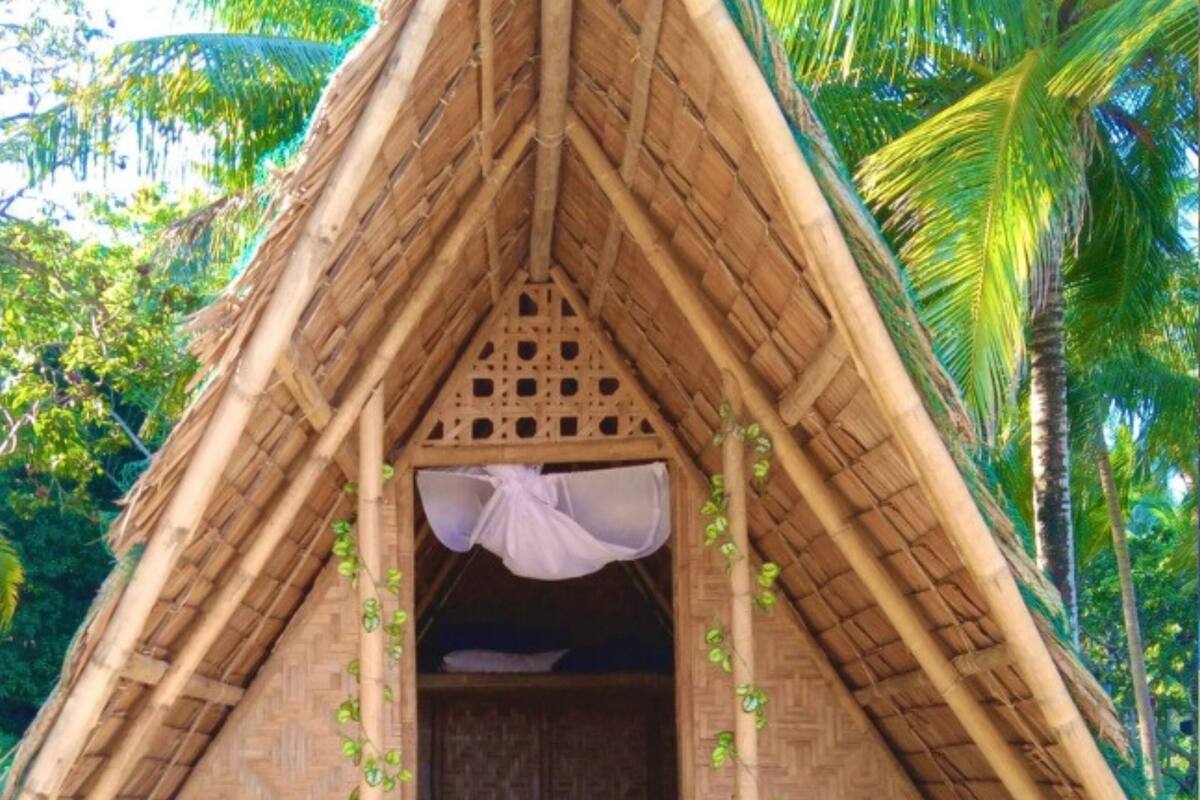 Eine strohgedeckte Hütte mit Bett und einer kleinen Veranda, umgeben von viel Grün.
