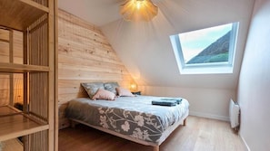 A bedroom with a sloped ceiling, a bed with floral bedding, a small window with a mountain view, and a wooden wall.
