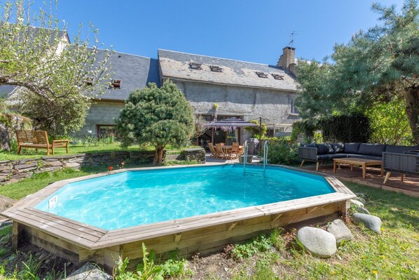 Ein Schwimmbad mit Holzterrasse, umgeben von einer Steinmauer, einer Terrasse mit Gartenmöbeln und einem Haus mit Schieferdach.