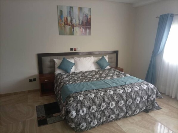 A neatly arranged bedroom with a large bed, a nightstand, a wall-mounted painting, and a window with curtains.