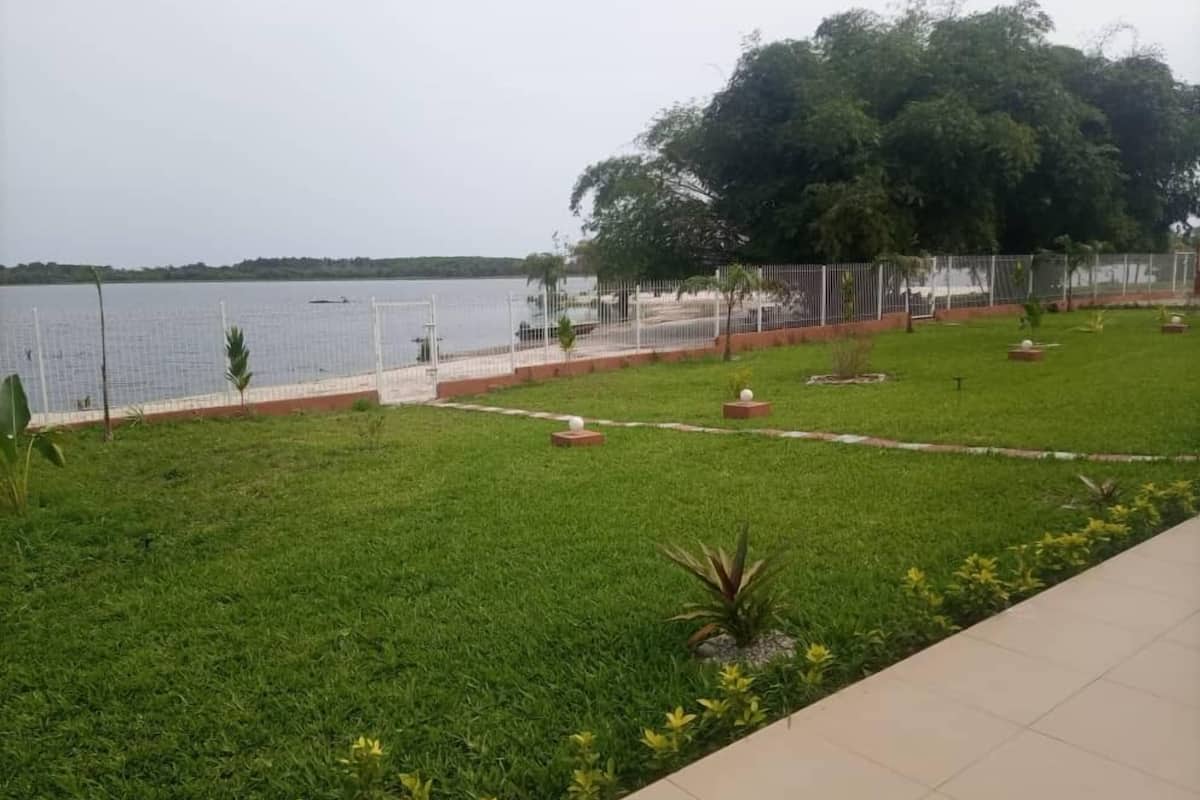 A grassy area with a view of a body of water, a fenced pathway, and a large tree.